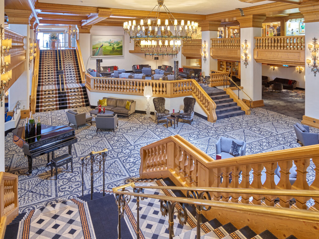 Die Hotelhalle über mehrere Ebenen: geschnitzte Holzbrüstungen, ein großer Kronleuchter, ein Flügel und gemusterte Böden
