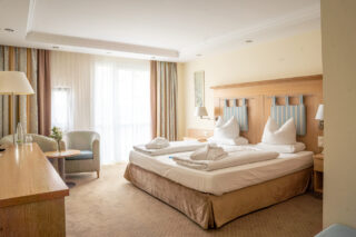 Helles Hotelzimmer mit Doppelbett, gemütlicher Sitzgelegenheit und einem großen Fenster, das viel Tageslicht hereinlässt.