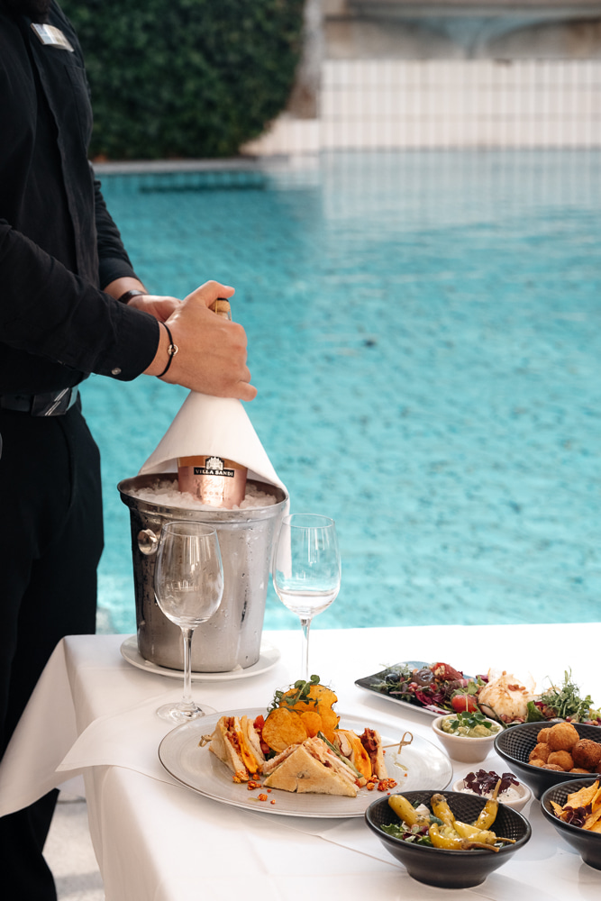 Kellner öffnet eine Flasche Champagner neben einem Tisch mit leckeren Snacks und frischem Salat am Pool.