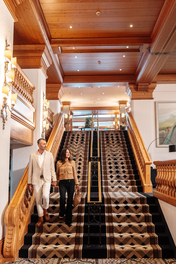 Paar geht eine elegante Treppe im Hotel hinauf, umgeben von luxuriösem Holzinterieur und stilvollen Lampen.