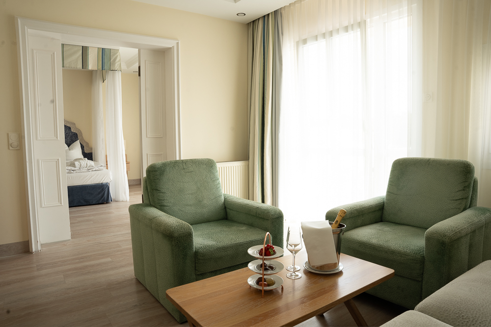 Hotelzimmer mit einem gemütlichen Sitzbereich, Blick auf das Bett und einem Tablett mit Snacks, Champagner und frischen Erdbeeren.