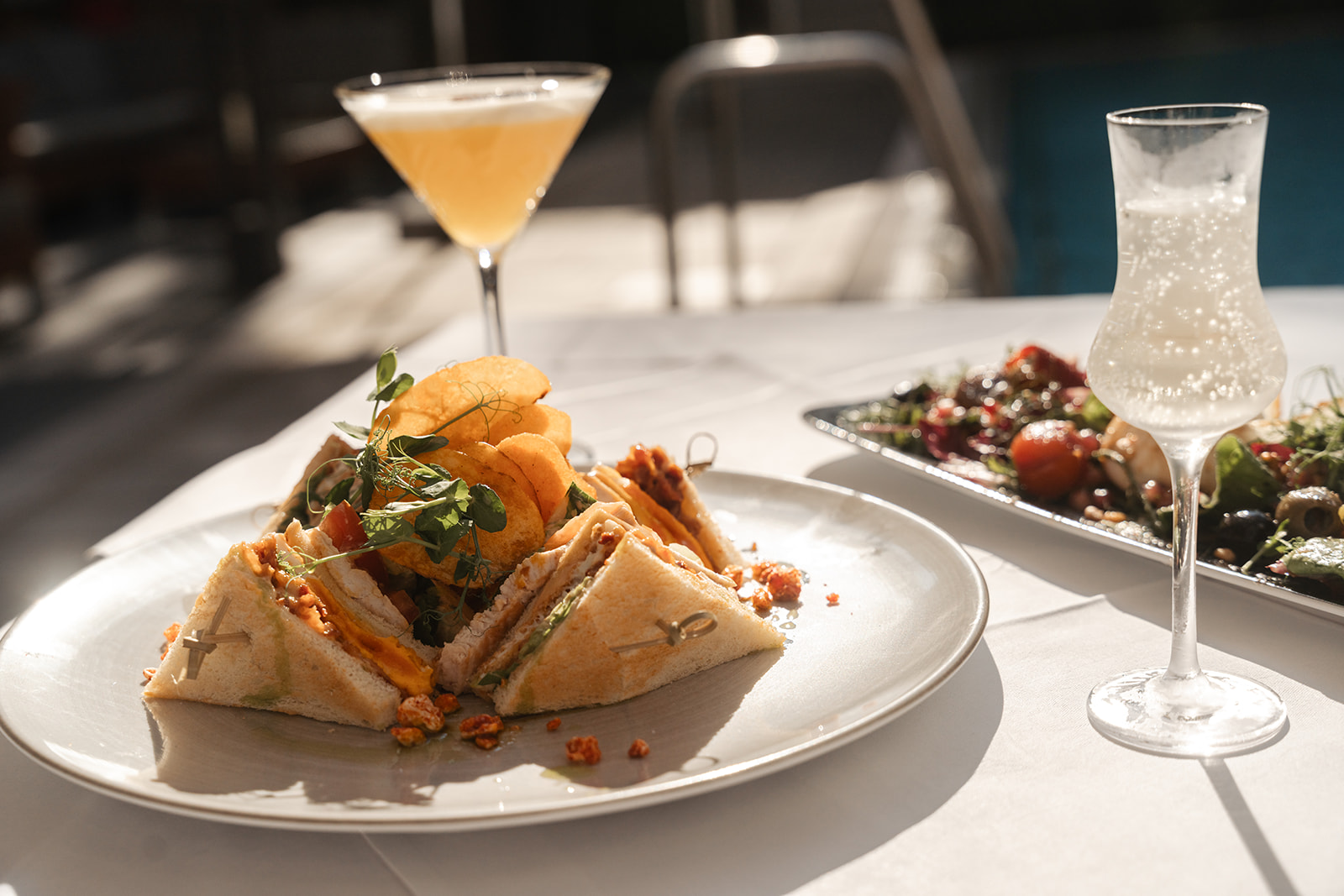 Sandwich mit Chips, frischen Kräutern und einem Cocktailglas auf einem Tisch, begleitet von einem frischen Salat.
