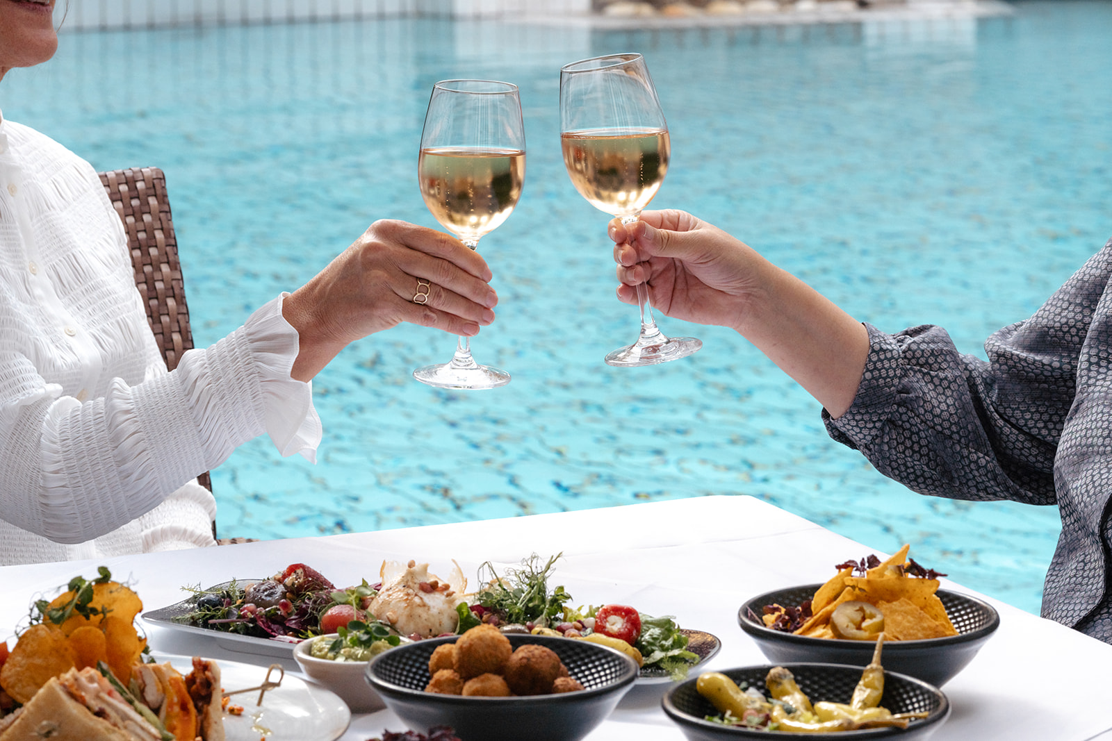 Zwei Personen stoßen mit Weingläsern an und genießen ein leckeres Mittagessen mit Salat und Snacks, während sie am Pool sitzen.