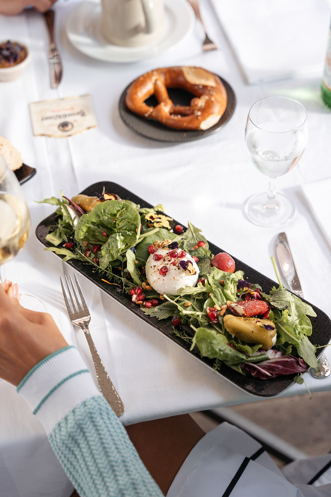 Frischer Salat mit Granatapfelkernen, Avocado und Mozzarella auf einem Teller, serviert mit einer Brezel und einem Glas Wasser.